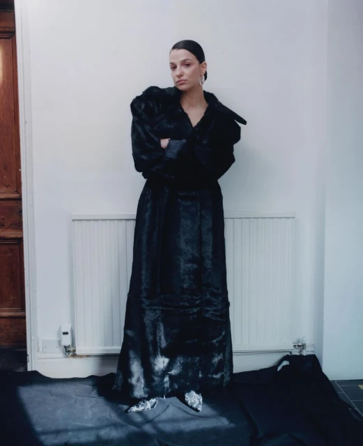 Woman in a black fur coat stands in front of a white wall, posing with her arms crossed in an indoor setting. Fashion photography.
