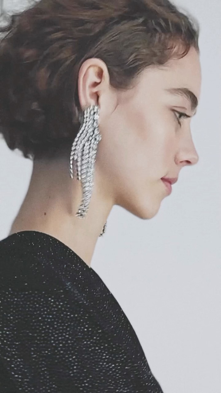 Profile portrait of a woman wearing large crystal fringe earrings and a black textured top against a plain light background.