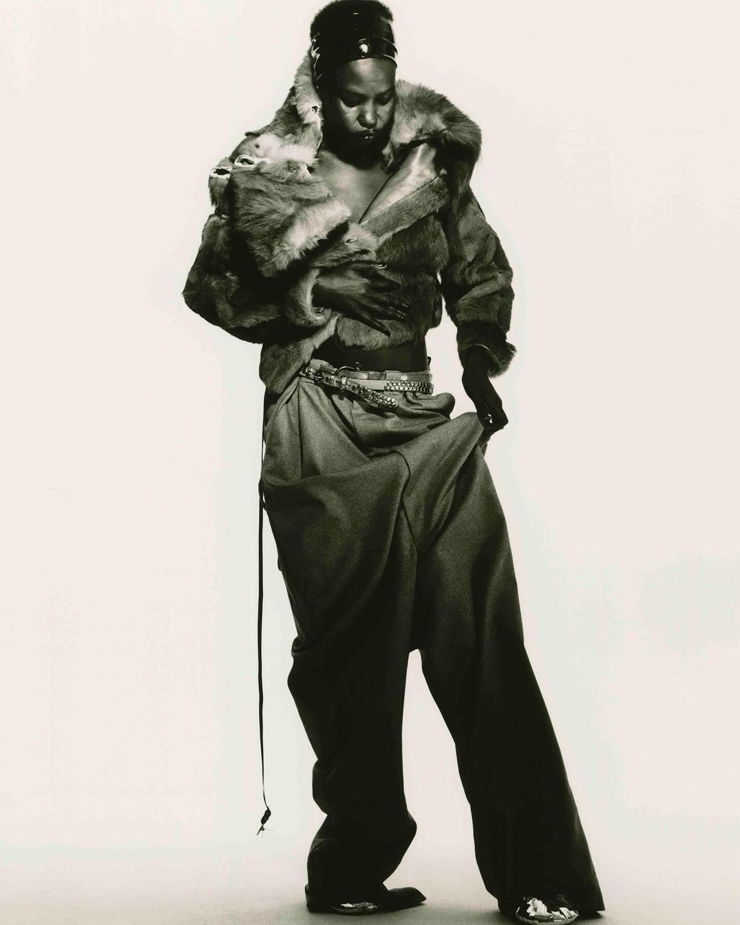 Black and white fashion portrait of an African American female model wearing a fur coat and baggy pants, with a light background.