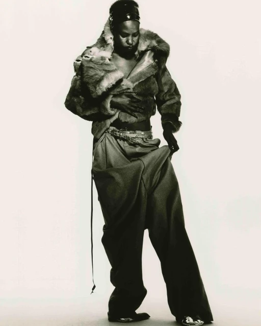 Black and white fashion portrait of an African American female model wearing a fur coat and baggy pants, with a light background.