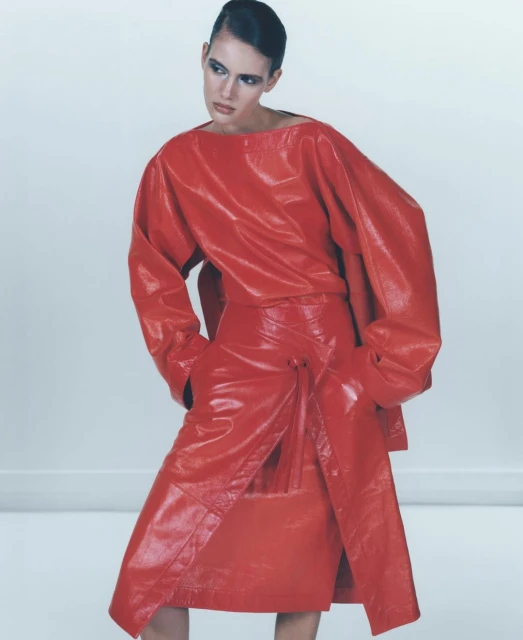 Woman in red patent leather outfit poses against white backdrop in a fashion shot, showcasing bold makeup and sleek hairstyle.