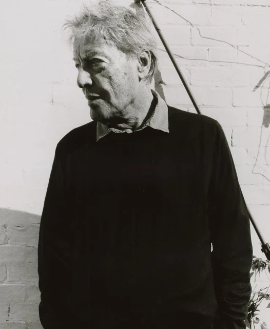 Black-and-white portrait of an elderly man with gray hair wearing a dark sweater, captured in natural light against a brick wall.