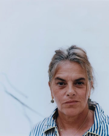 Portrait of an older woman with grey-streaked hair wearing a striped collared shirt against a plain light wall in London.