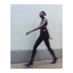 Motion-blurred profile of a model walking in dark clothing against a light wall by Charlie Gates