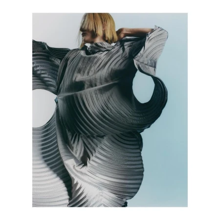 Model with blonde bob wearing a pleated grey sculptural dress against a gradient background by Charlie Gates.