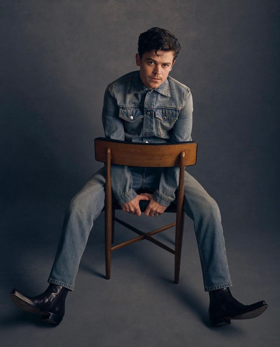 Man in denim jacket and jeans seated backwards on wooden chair against dark grey backdrop