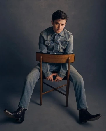 Man in denim jacket and jeans seated backwards on wooden chair against dark grey backdrop