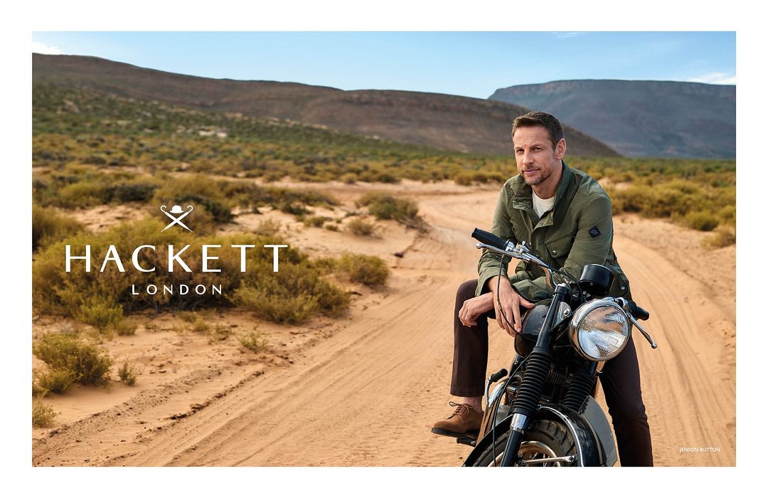 An adult male wearing a green jacket and brown pants is sitting on a motorcycle on a dirt road with desert hills in the background.