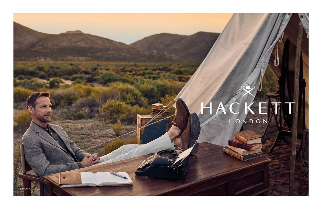 Man in a blazer sits at an outdoor desk with a typewriter and books, near a tent in a desert landscape with mountains, Hackett London ad.