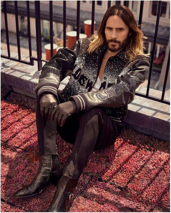 Jared Leto poses on a rooftop, wearing a black patterned jacket, dark pants, and boots, sitting on red textured surface with city views.
