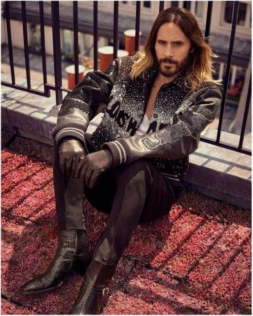 Jared Leto poses on a rooftop, wearing a black patterned jacket, dark pants, and boots, sitting on red textured surface with city views.