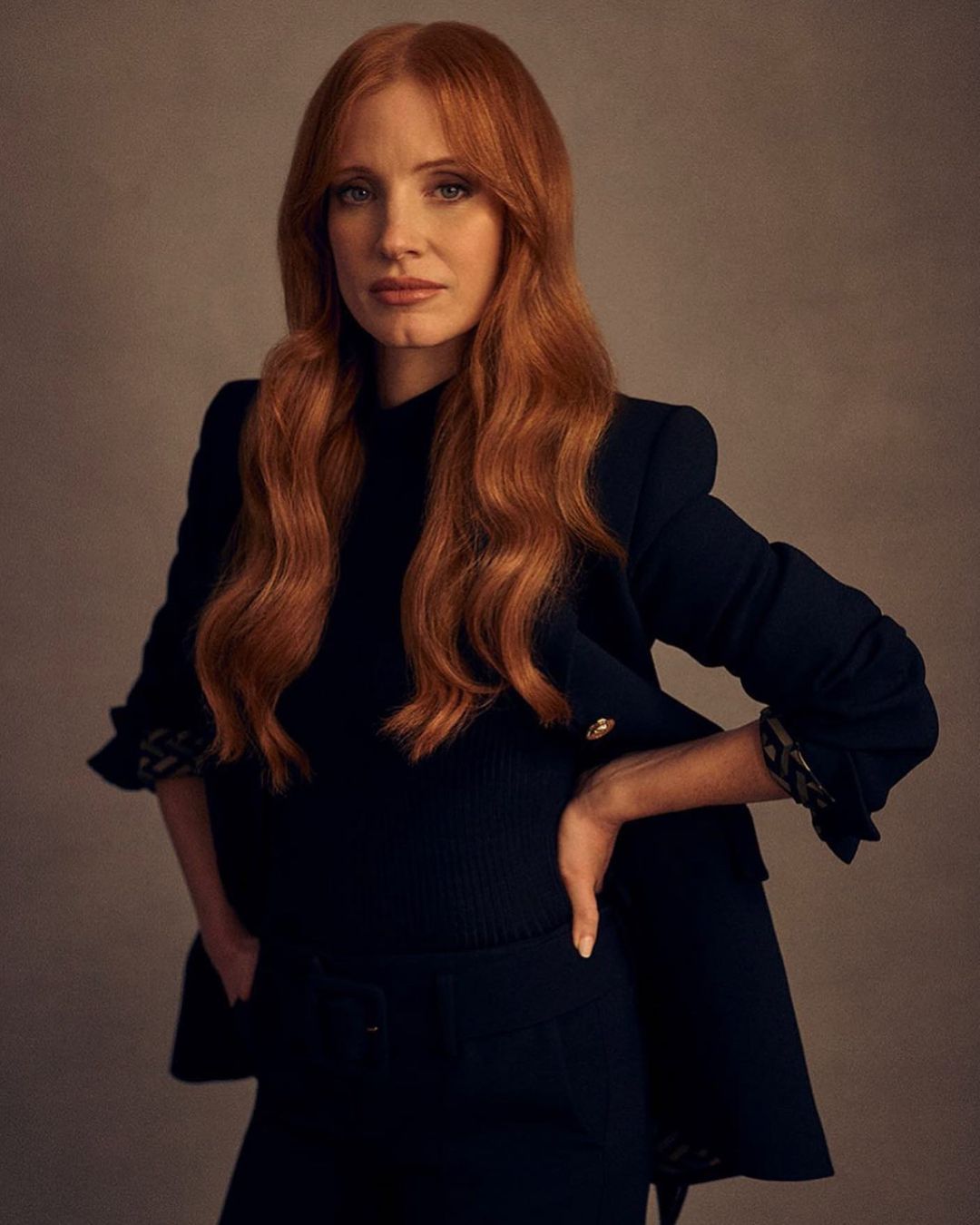 Jessica Chastain poses in a dark blazer and matching pants in a portrait, showcasing her long red hair and stylish attire.