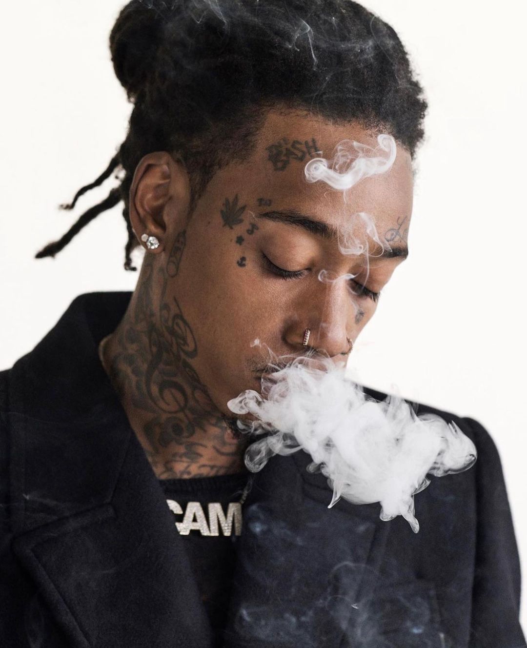 Portrait of a tattooed man with dreadlocks exhaling a cloud of smoke against a plain white background in a close-up shot.