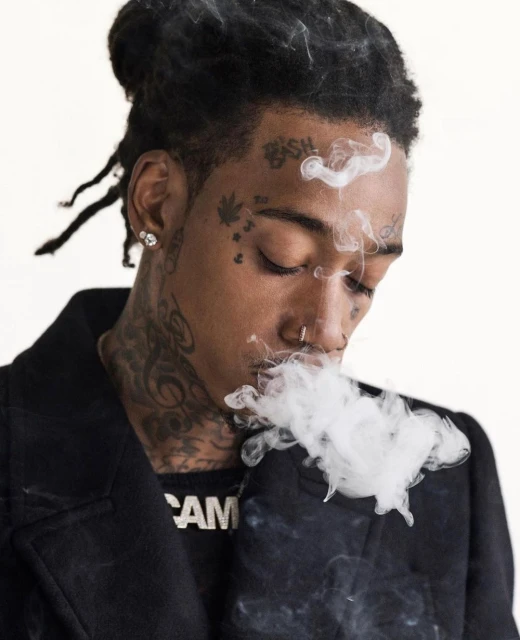 Portrait of a tattooed man with dreadlocks exhaling a cloud of smoke against a plain white background in a close-up shot.