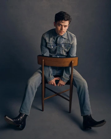 Man wearing a denim jacket and jeans straddling a wooden chair in front of a dark textured studio backdrop