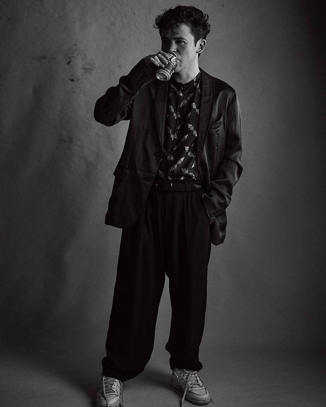 Black and white portrait of a young man drinking from a beverage can, wearing a patterned shirt, blazer, and casual shoes.