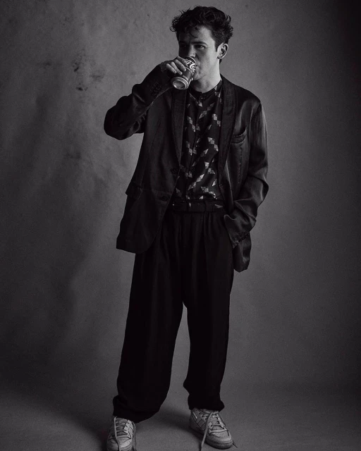 Black and white portrait of a young man drinking from a beverage can, wearing a patterned shirt, blazer, and casual shoes.