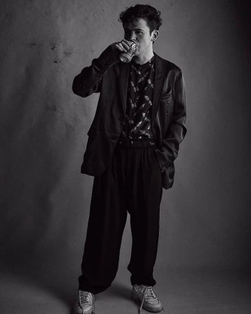 Black and white portrait of a young man drinking from a beverage can, wearing a patterned shirt, blazer, and casual shoes.