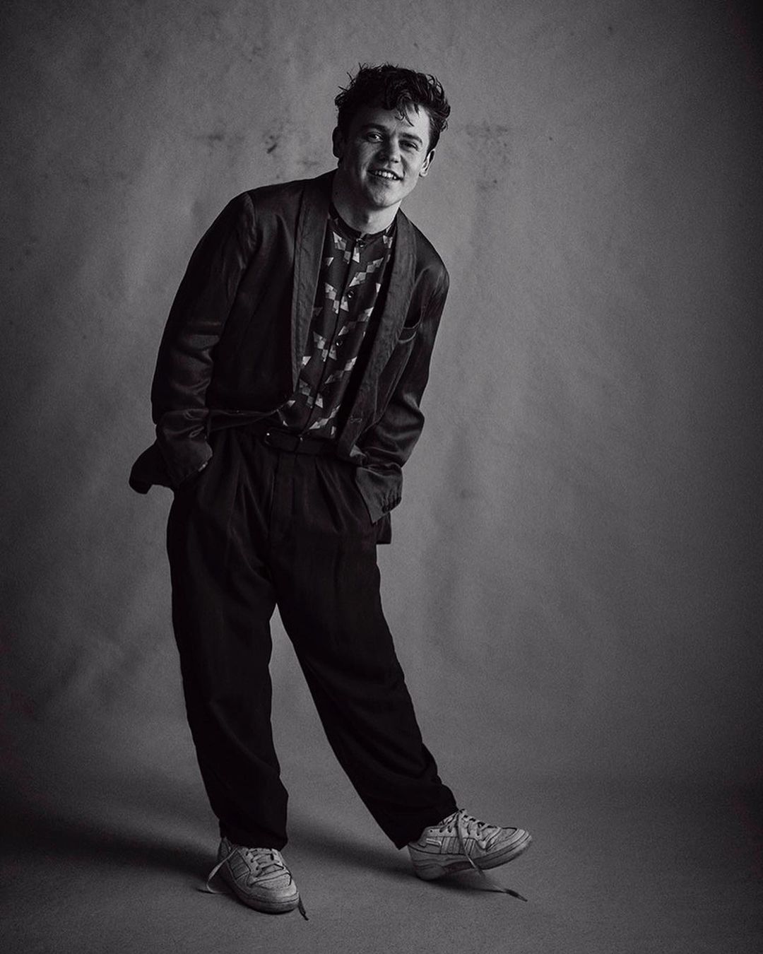 Monochrome portrait of smiling young man in blazer and trousers standing against textured studio backdrop
