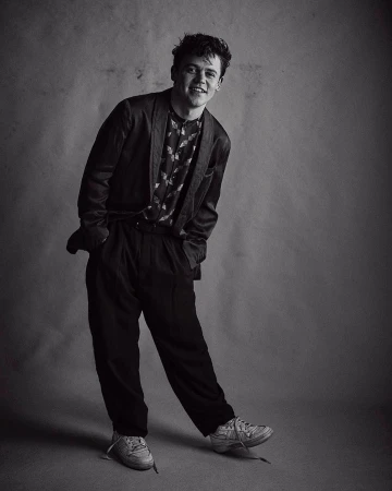 Monochrome portrait of smiling young man in blazer and trousers standing against textured studio backdrop