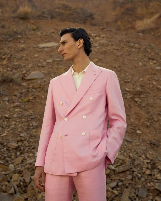A man in a pink suit stands against a rocky, mountainous backdrop, looking away from the camera in an outdoor fashion shot.