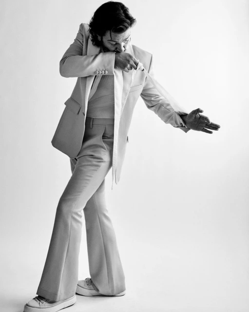 Monochrome portrait of a man in a suit jacket and bell-bottom pants, striking a pose in front of a white backdrop. Studio lighting.