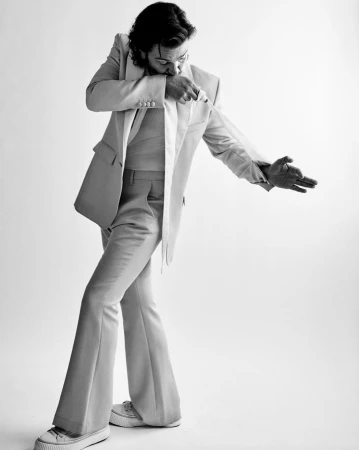 Monochrome portrait of a man in a suit jacket and bell-bottom pants, striking a pose in front of a white backdrop. Studio lighting.