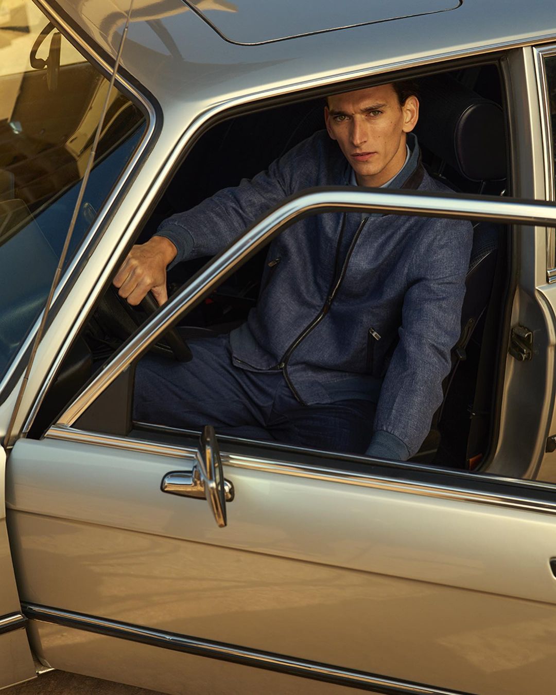 Male model in blue denim jacket poses inside a vintage silver car with open door. Automotive fashion editorial shot.