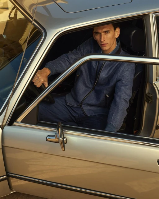Male model in blue denim jacket poses inside a vintage silver car with open door. Automotive fashion editorial shot.