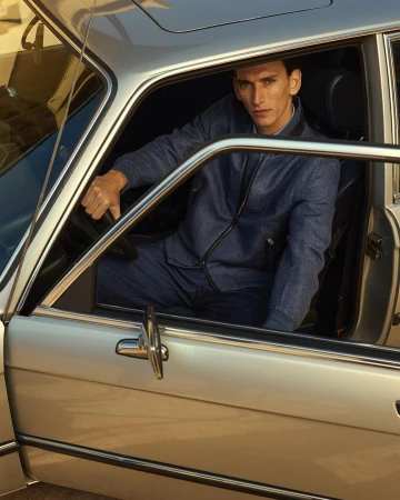 Male model in blue denim jacket poses inside a vintage silver car with open door. Automotive fashion editorial shot.