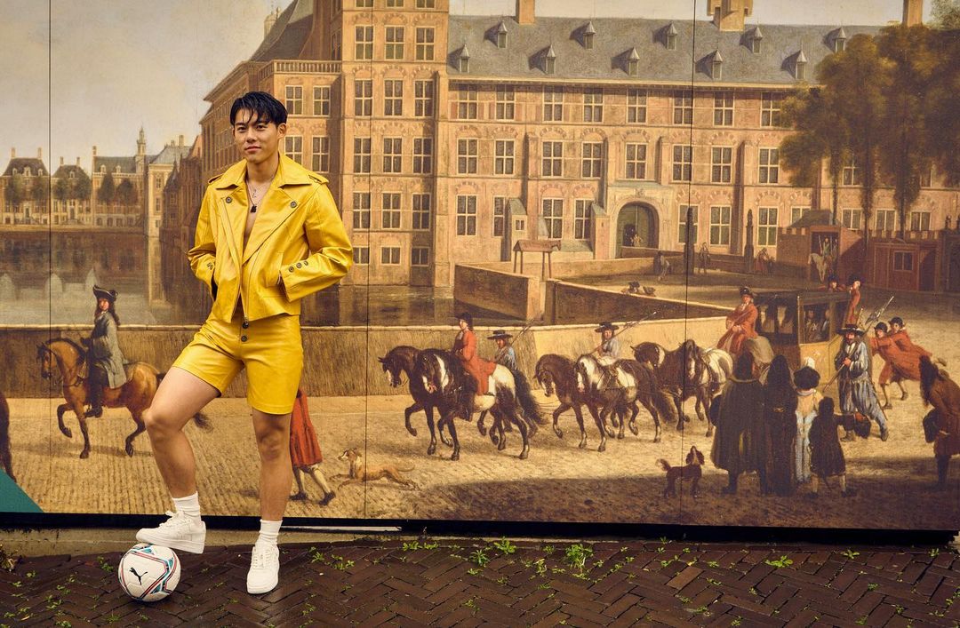 Asian man in a yellow jacket and shorts poses with a soccer ball in front of a large mural in an urban environment on a brick surface.