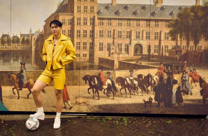 Asian man in a yellow jacket and shorts poses with a soccer ball in front of a large mural in an urban environment on a brick surface.