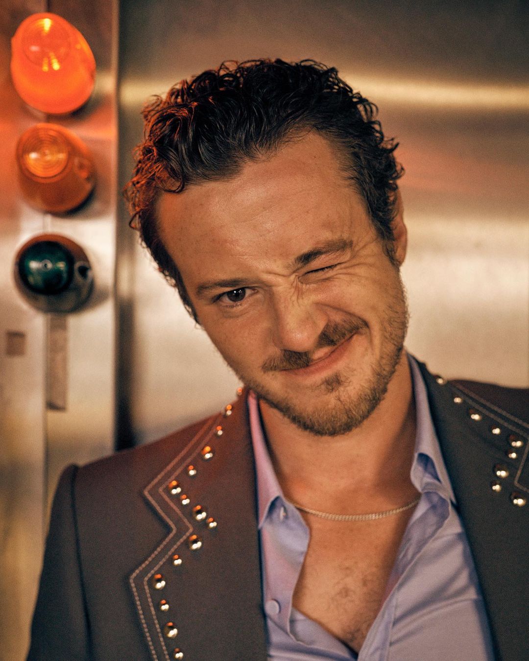 Portrait of adult man with curly brown hair winking while wearing a blazer with studs on a neutral background.