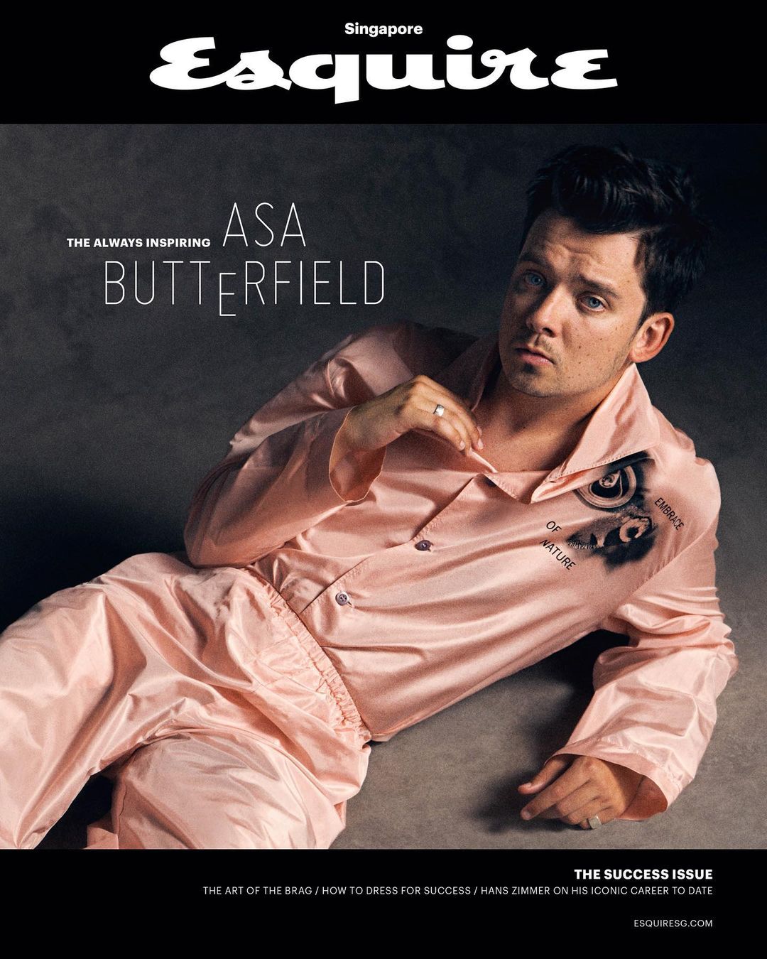 Asa Butterfield poses on an Esquire Singapore cover. He's lying in a pink pajama set against a dark gray background. He gazes directly at the viewer.