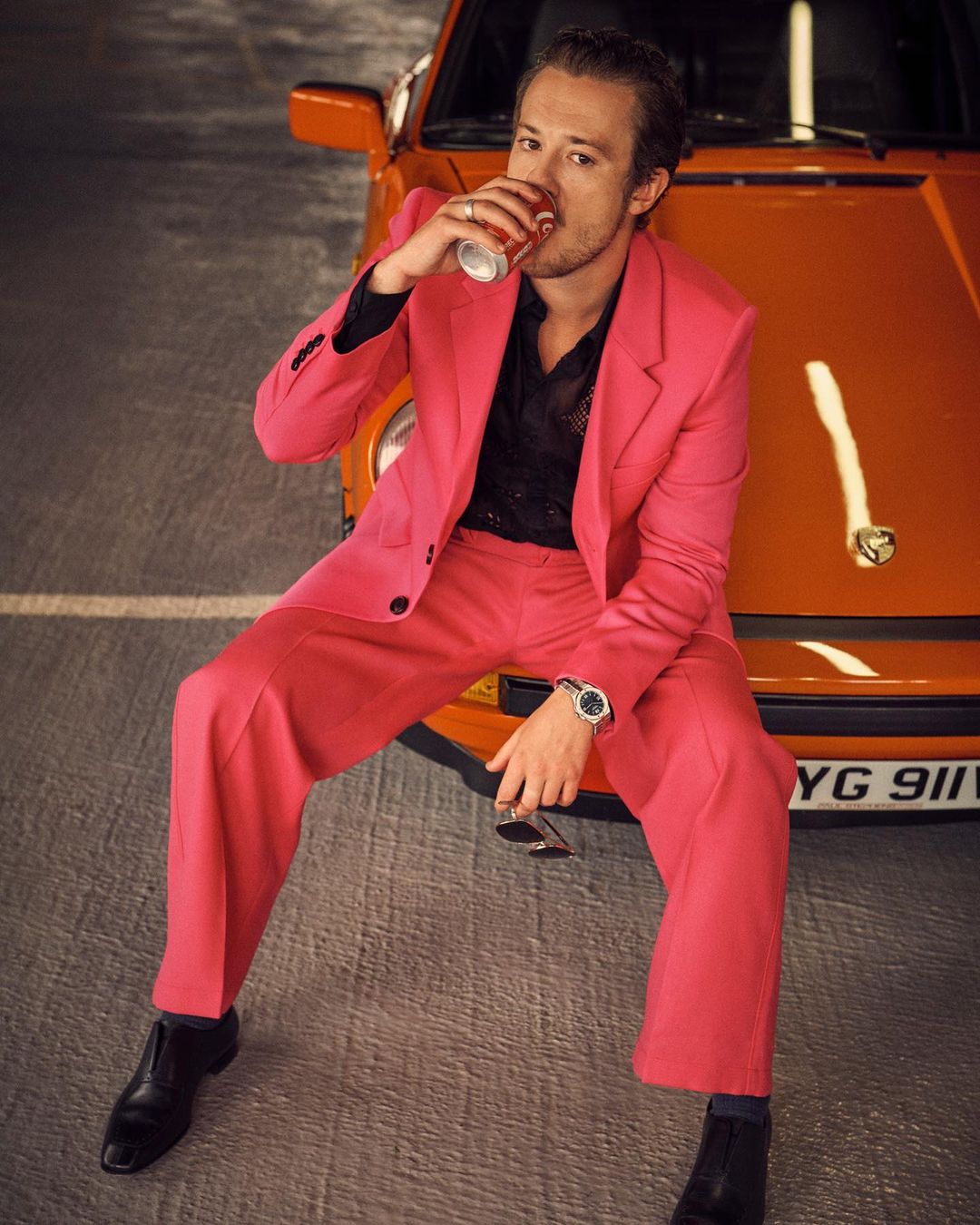 Man in a pink suit seated on an orange car drinks from a can, showcasing retro style and automotive fashion against a concrete backdrop.