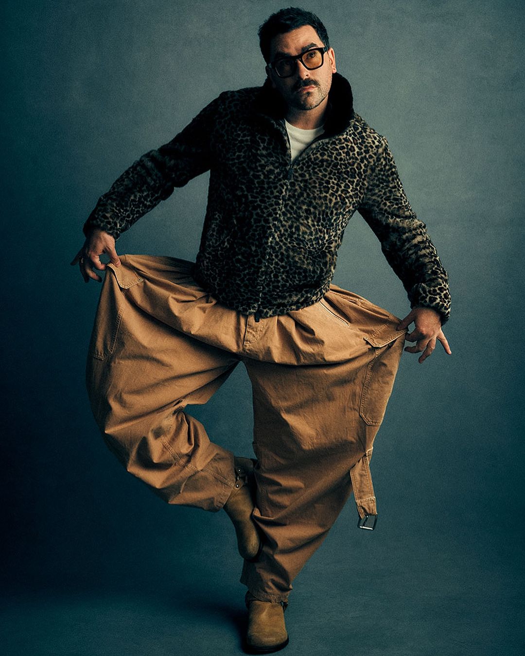 A man in a patterned jacket and tan pants poses dynamically against a grey background, showcasing a unique style and confident stance.