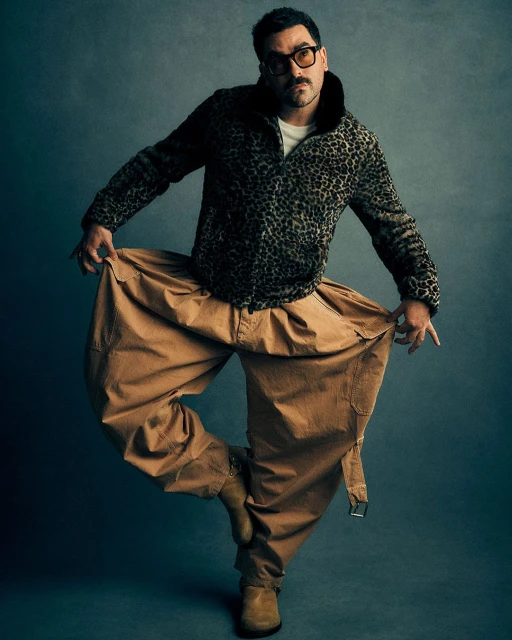 A man in a patterned jacket and tan pants poses dynamically against a grey background, showcasing a unique style and confident stance.