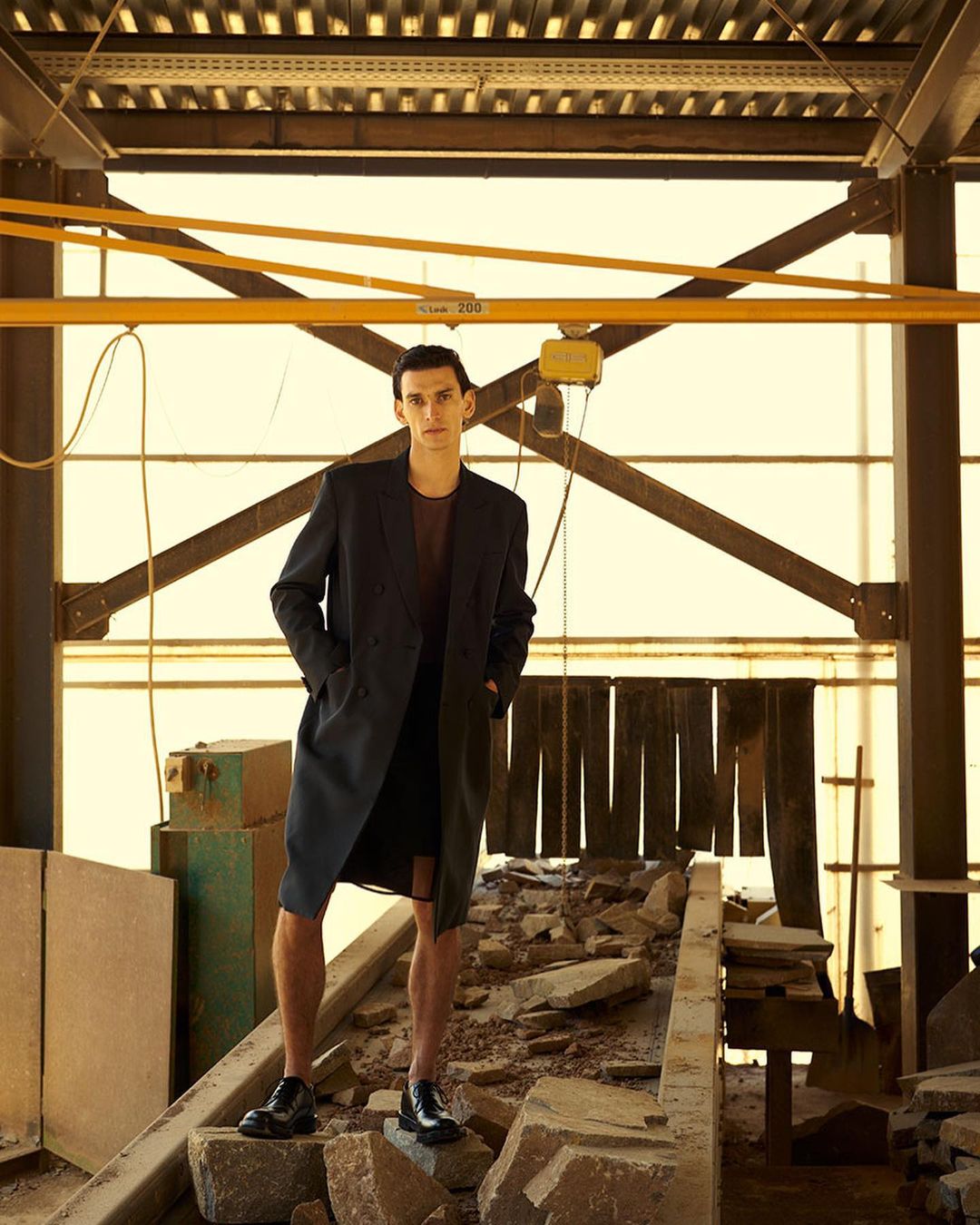 A tall man in a black coat poses on stone rubble in an industrial setting with yellow overhead beams. Fashion editorial shot.