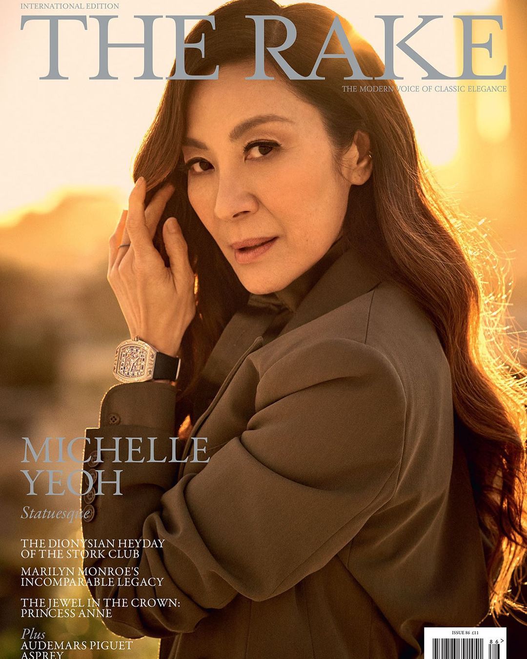 Michelle Yeoh graces The Rake magazine cover in a medium shot. She wears a sophisticated coat and jewelry, with a blurred city view.