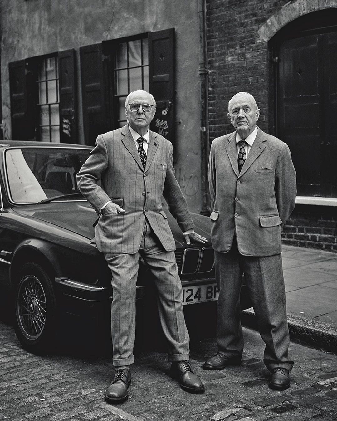 Two older men in suits stand beside a vintage car on a cobblestone street near a brick building with window shutters in a black-and-white photo.