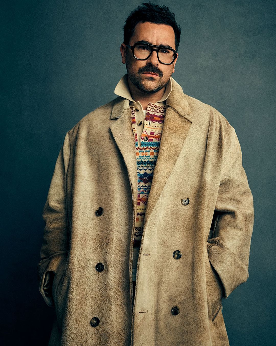 Headshot of a stylish adult man wearing a tan overcoat, eyeglasses, and patterned sweater against a dark gray studio backdrop.