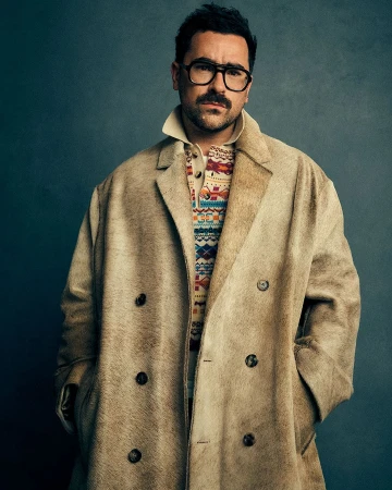 Headshot of a stylish adult man wearing a tan overcoat, eyeglasses, and patterned sweater against a dark gray studio backdrop.