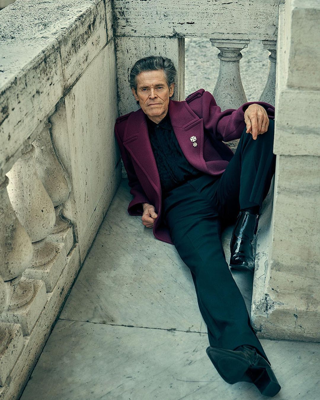 An adult male is lying on the marble floor and leaning against a marble ledge wearing a purple coat and black pants and boots.