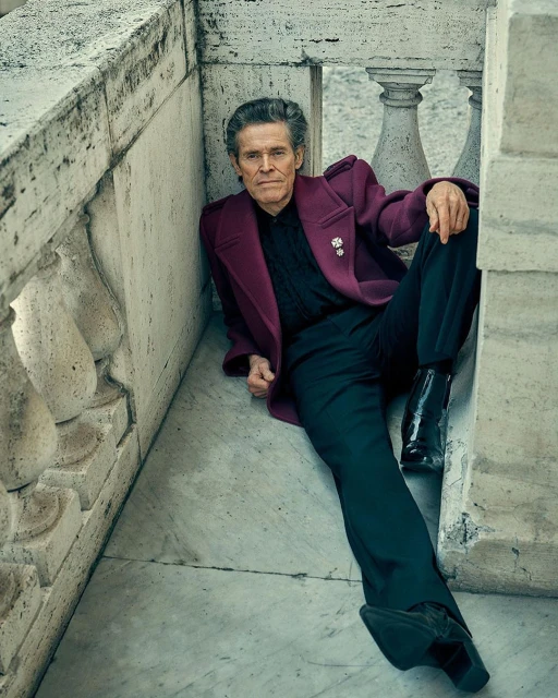 An adult male is lying on the marble floor and leaning against a marble ledge wearing a purple coat and black pants and boots.