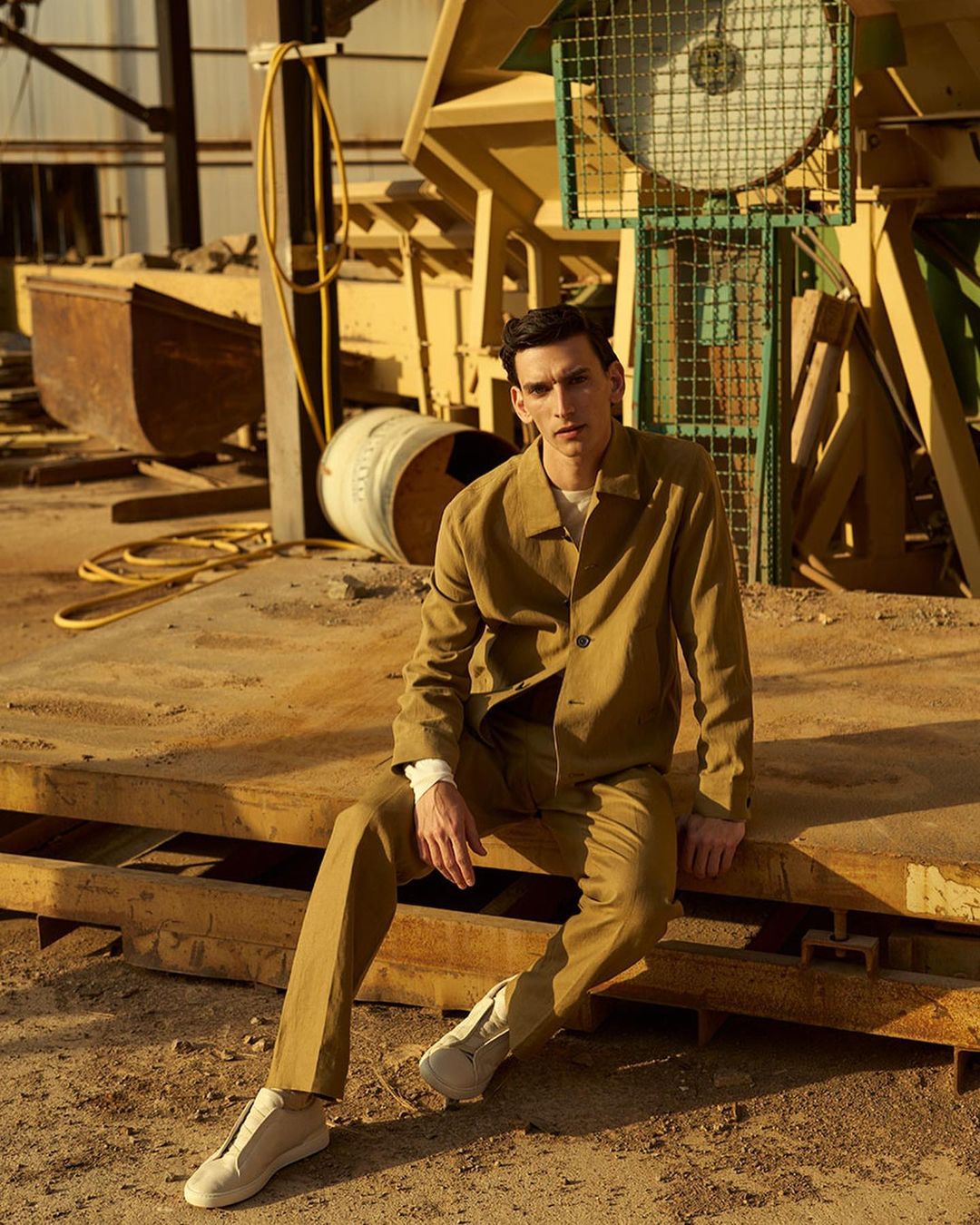 A young adult male wearing a khaki suit sits on a yellow surface in an industrial setting on a sunny day, looking at the camera.
