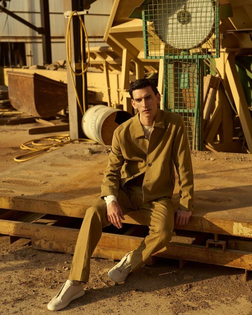 A young adult male wearing a khaki suit sits on a yellow surface in an industrial setting on a sunny day, looking at the camera.