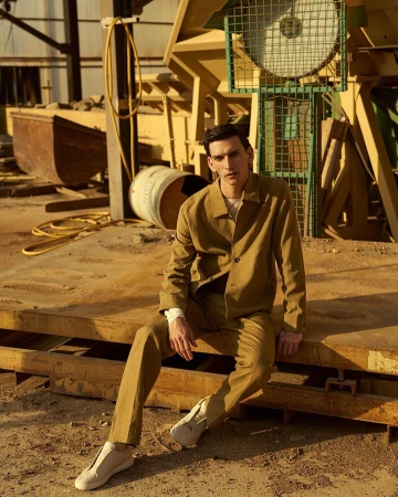 A young adult male wearing a khaki suit sits on a yellow surface in an industrial setting on a sunny day, looking at the camera.