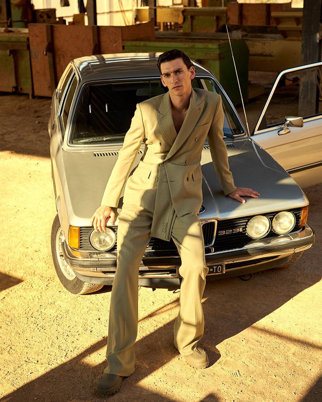 A stylish adult male model leans on a silver vintage car outdoors in a tan suit, creating a striking fashion image.
