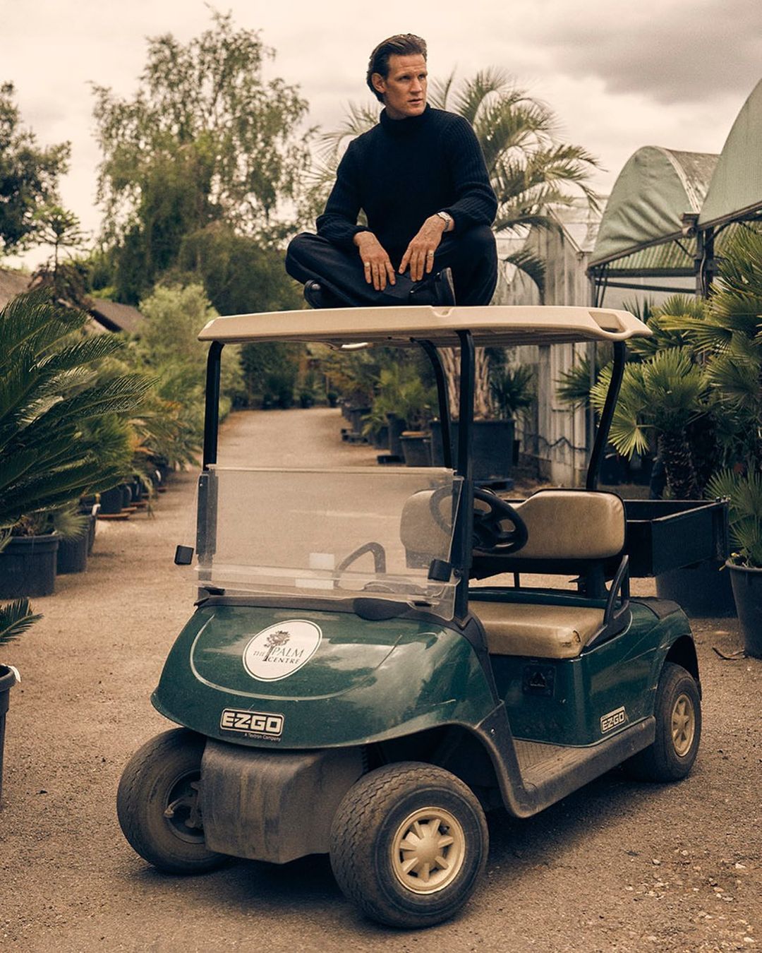 Matt Smith sits cross-legged atop a green golf cart in a garden centre, surrounded by potted palm trees and foliage, under soft daylight.