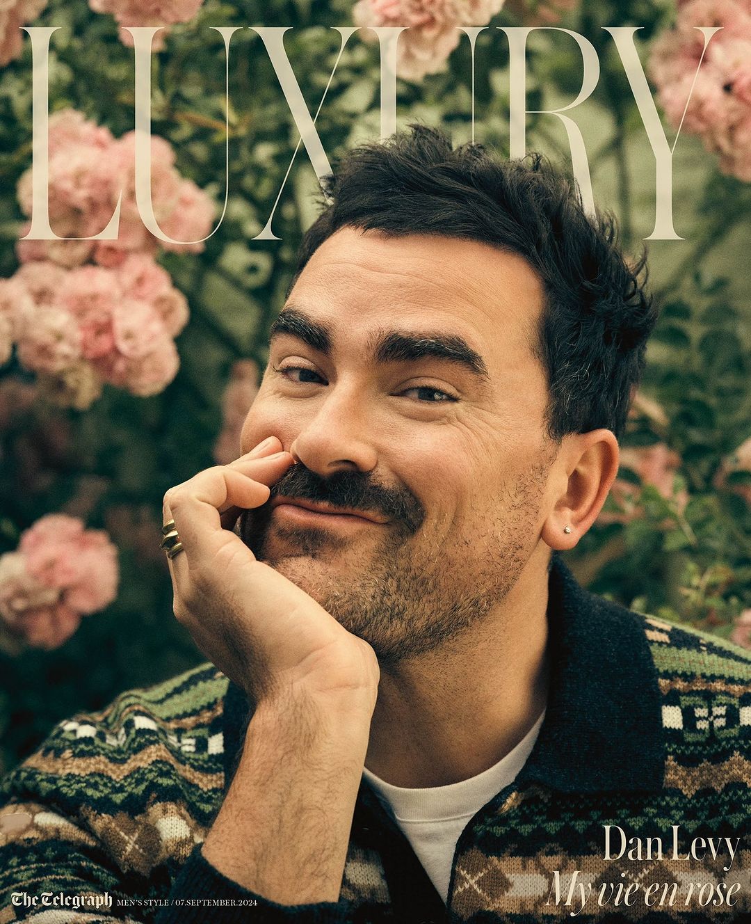 Dan Levy poses in a magazine cover portrait wearing a patterned sweater with pink flowers in the background and the word 'luxury' above him.
